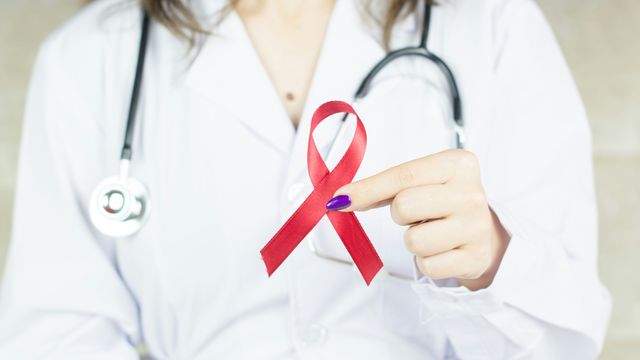 A female doctor in a white lab coat holding up a red ribbon representing HIV awareness. 
