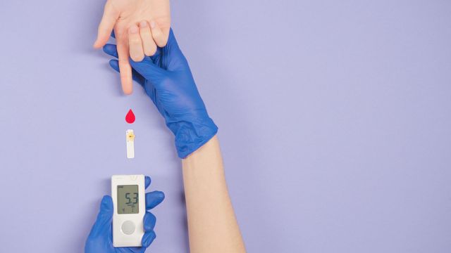 Medical professional wearing blue gloves holds a blood testing machine and a person's hand with finger outstretched. 