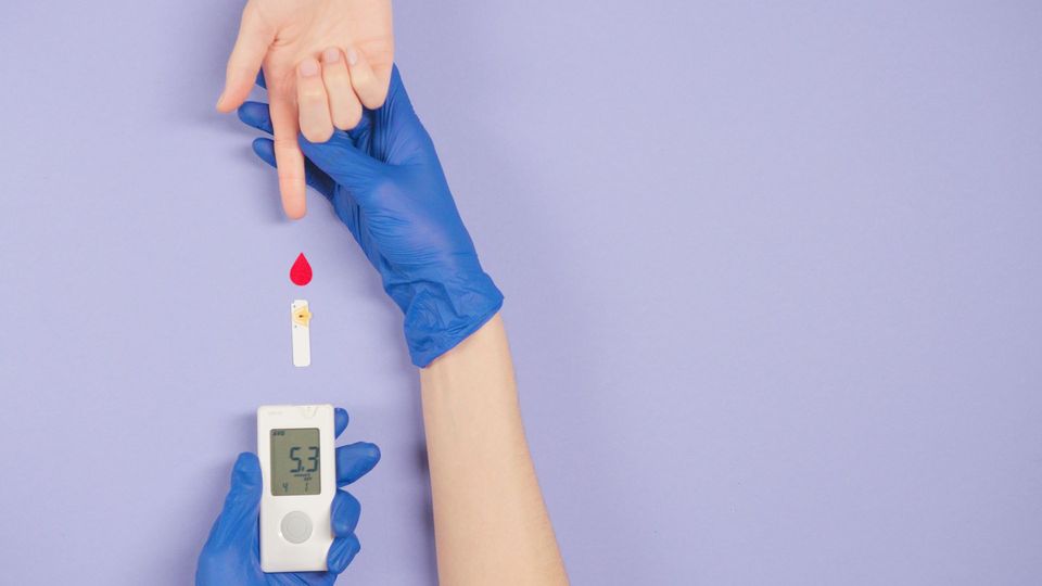 Medical professional wearing blue gloves holds a blood testing machine and a person's hand with finger outstretched.