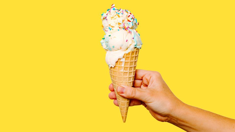 Hand holding a waffle cone with colorful scoops of ice cream and rainbow sprinkles.