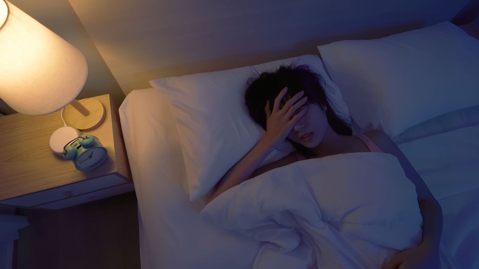 Person lying in bed under white covers, hand across face, in dim blue lighting, representing sleep.