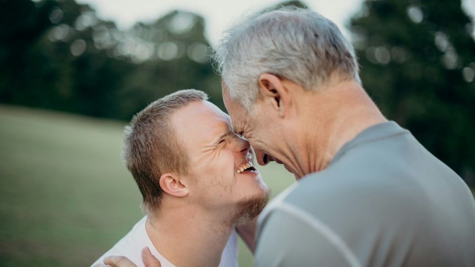 Young person with Down syndrome sharing a joyful embrace outdoors with an older adult in a grassy park.