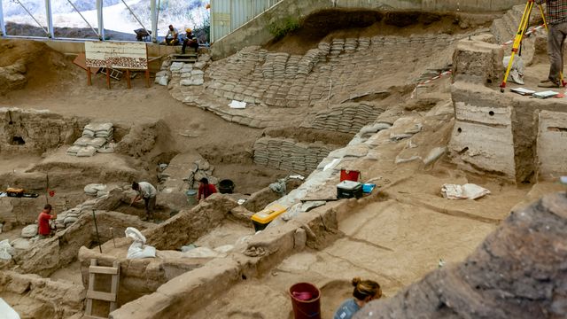 Archaeology excavation site with exposed ancient structures, trenches, and researchers documenting findings. 