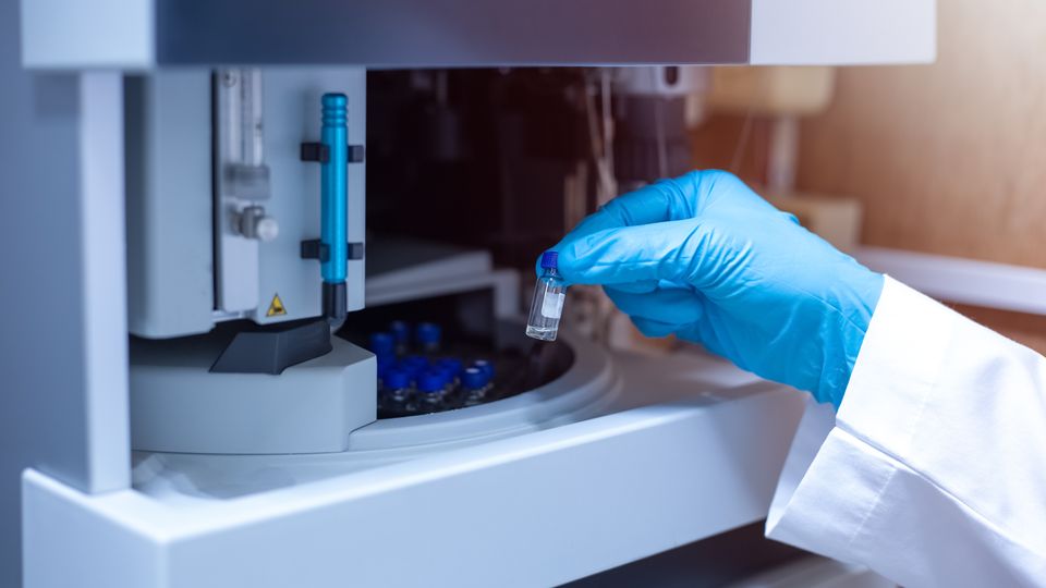 Gloved hand placing a sample vial into an LC laboratory autosampler for automated analysis.