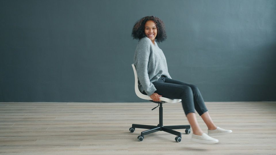 Person seated upright on a wheeled office chair with feet extended forward, demonstrating seated posture.