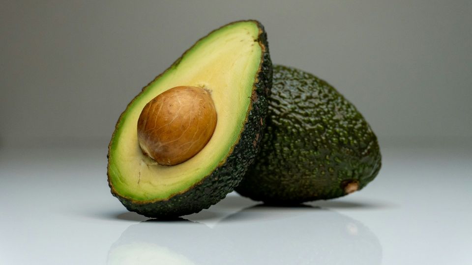 Ripe avocado cut in half with the pit intact beside a whole avocado on a white background.