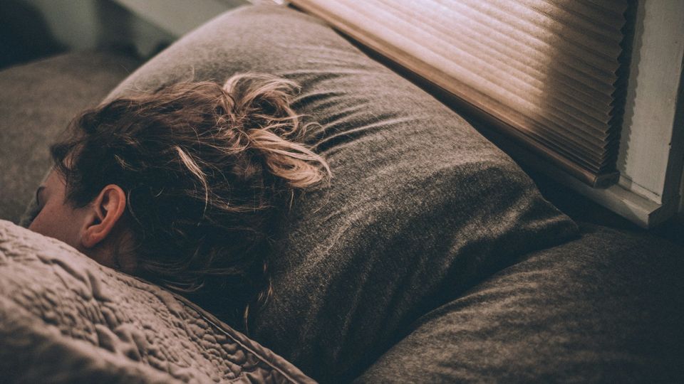 Woman sleeping peacefully on a couch under a blanket beside a window.