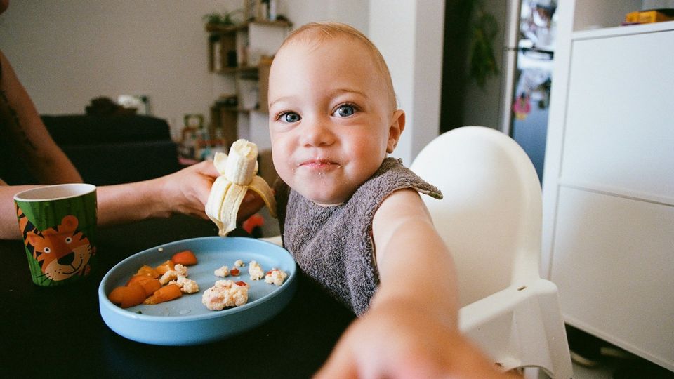 Baby eating a banana in a high chair during mealtime with healthy finger foods on a plate.