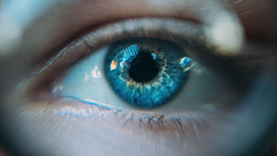 Blue eye in close-up macro view, showing detailed iris patterns, eyelashes, and light reflections.