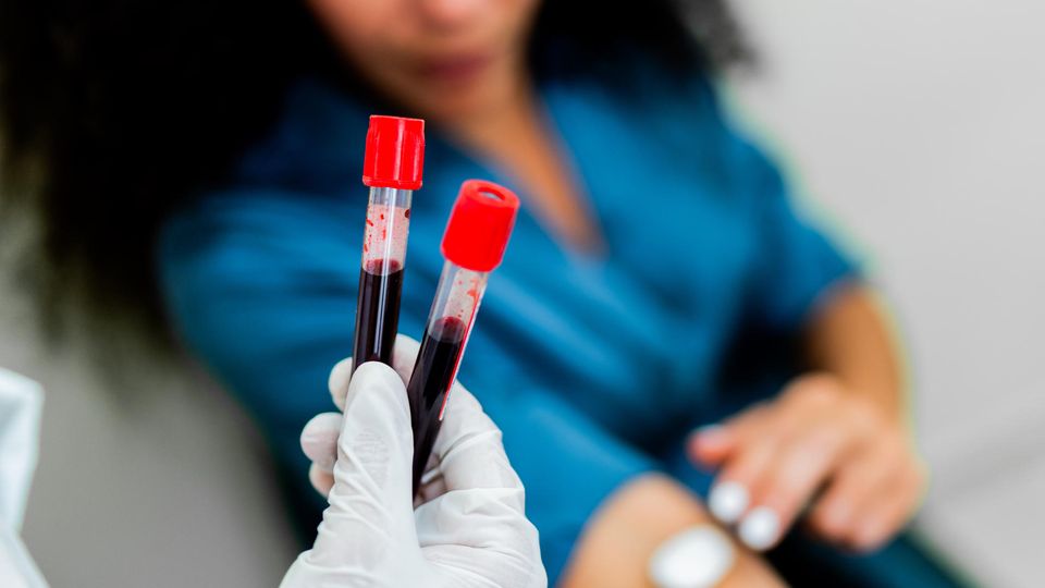 Gloved hand holding two blood-filled test tubes, with a woman holding a cotton pad on her arm in the background.