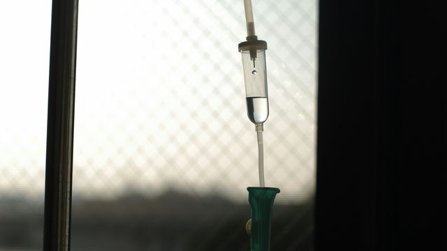 Close-up of a hospital drip set-up in a dark room, with light coming through a window in the background. 