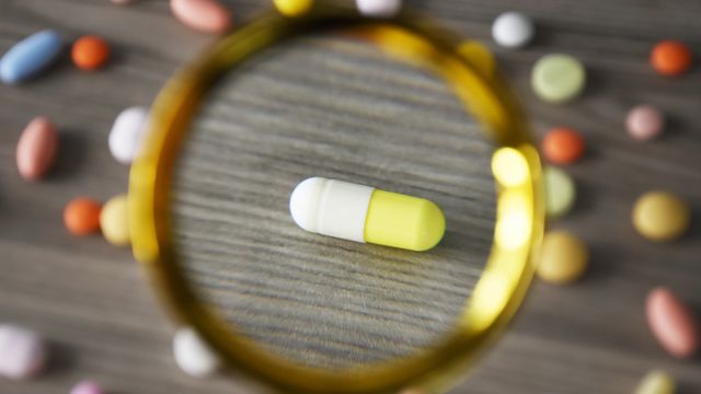 Drug capsule viewed through a magnifying glass on a tabletop, surrounded by assorted colourful tablets. 