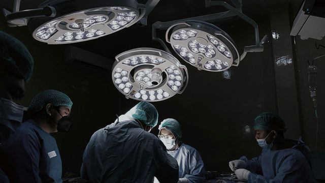 Surgeons wearing PPE under bright lights in an operating theatre, while the patient is under anesthesia. 