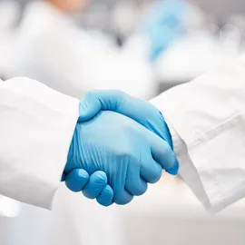 Two scientists shaking hands in white lab coat and blue rubber gloves.