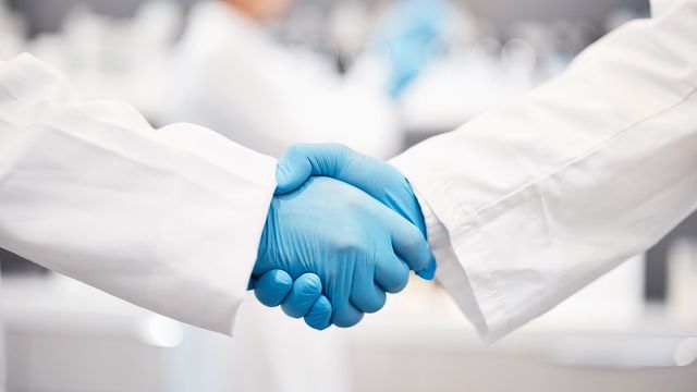 Two scientists shaking hands in white lab coat and blue rubber gloves. 