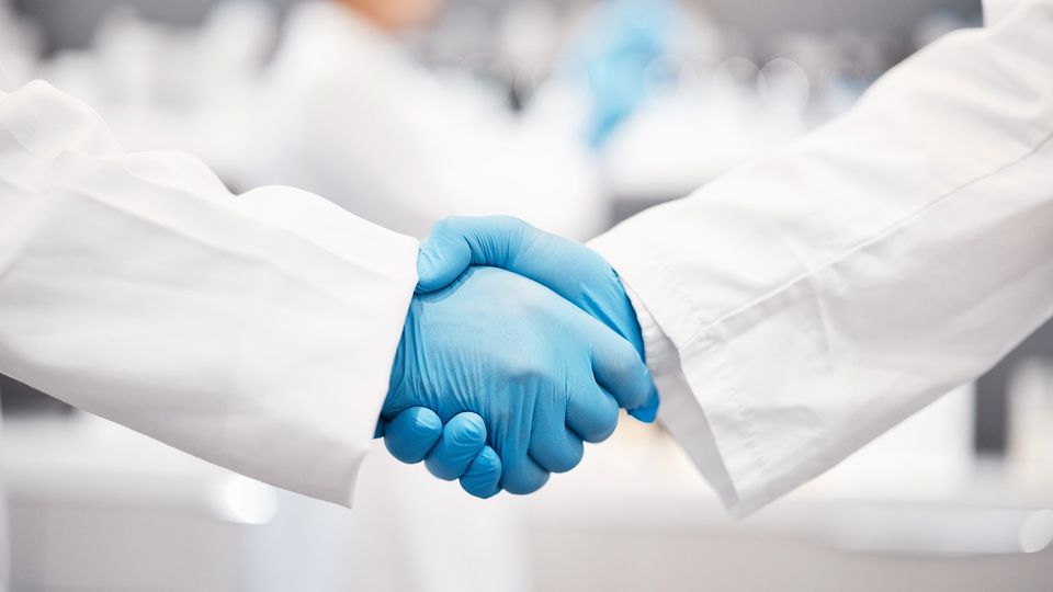 Two scientists shaking hands in white lab coat and blue rubber gloves.