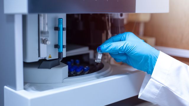 Gloved hand loading a sample vial into an automated LC-MS instrument in a laboratory. 