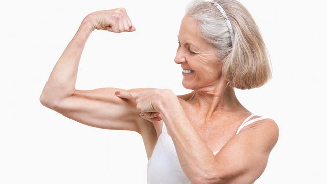 An older women pointing to her flexed bicep and smiling. 