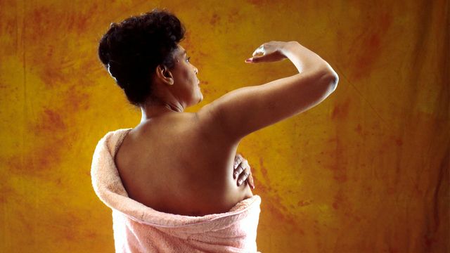 Woman performing a breast self-exam, checking her breast for changes, against an orange background. 