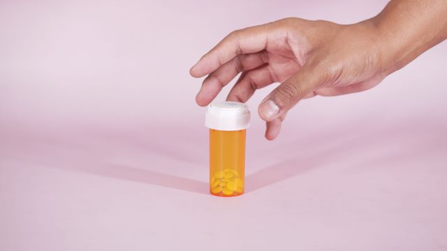 A hand reaching for a container filled with pills, which is placed on a pin surface. 