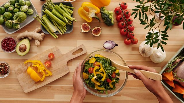 A kitchen worktop with different fruits and vegetables, as a person makes a salad in a bowl. 