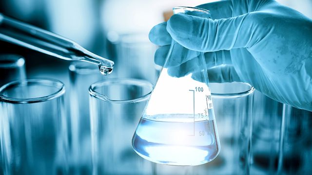 Gloved hand holding a laboratory flask while a pipette dispenses water, illustrating the role of water quality in chemical trace analysis. 