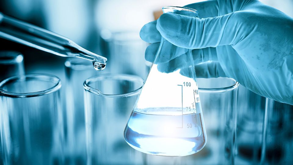Gloved hand holding a laboratory flask while a pipette dispenses water, illustrating the role of water quality in chemical trace analysis.