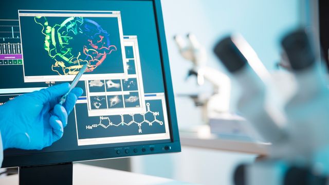 A gloved hand points at a computer screening showing a protein molecule with microscopes in the background. 