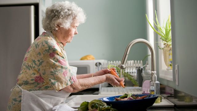 An older woman washing vegetables by a kitchen sink. 