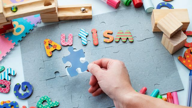 Hand placing puzzle piece beside colourful letters spelling autism, with toys and markers on a tabletop. 