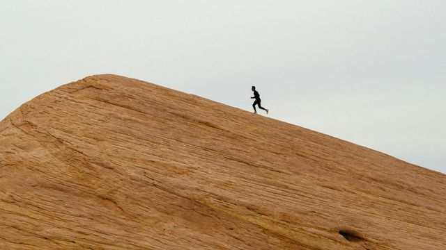 Far-away shot of a person running up a steep hill, with a gray sky in the background. 
