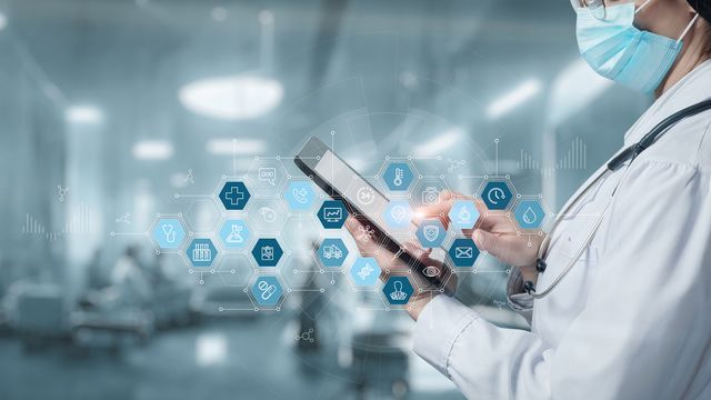 A doctor holding a tablet in a hospital ward behind an overlay of blue health care icons in hexagonal boxes.  