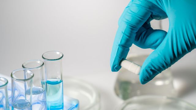 A capsule held by a gloved hand, next to test tubes containing blue liquid in a laboratory rack. 