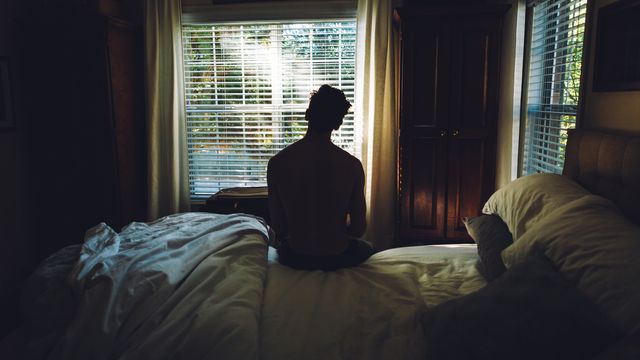Silhouette seated on an unmade bed facing a bright window, symbolising depression and isolation. 