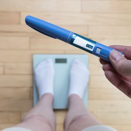 A person standing on a set of electronic scales while holding a GLP-1 "pen" injector. 