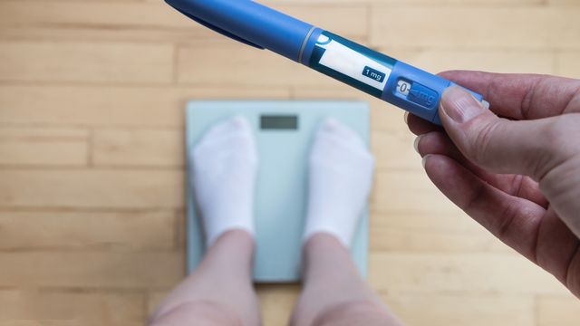 A person standing on a set of electronic scales while holding a GLP-1 "pen" injector. 
