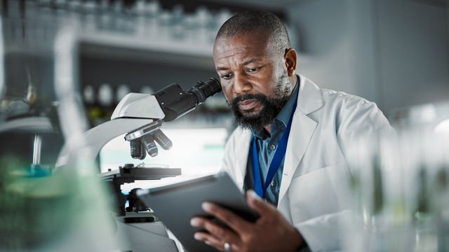 Male scientist seated next to a microscope holding a tablet.  