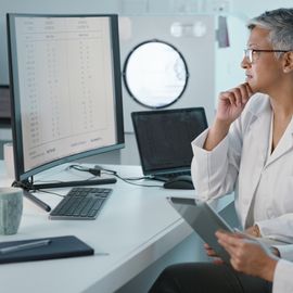 Two female scientists in white lab coats sitting looking at a computer screen displaying scientific data. 