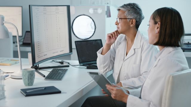 Two female scientists in white lab coats sitting looking at a computer screen displaying scientific data. 