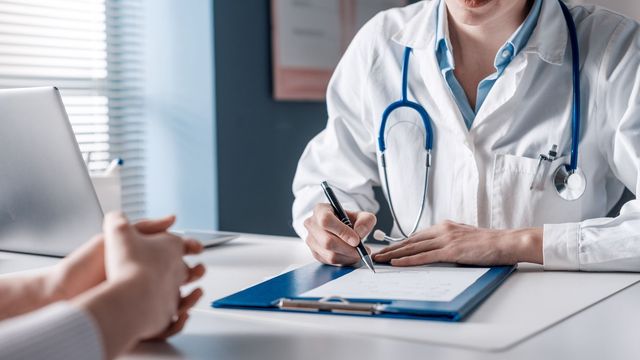 Doctor wearing a white coat and stethoscope, writing notes on a clipboard during a patient consultation. 