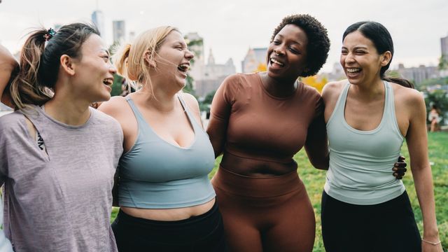 Women in athletic wear standing arm in arm outdoors in a park, gathered after a group fitness session. 