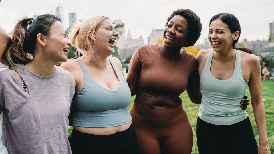 Women in athletic wear standing arm in arm outdoors in a park, gathered after a group fitness session.