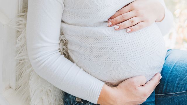 Pregnant person cradling their belly while seated, wearing a white knitted jumper and blue jeans. 