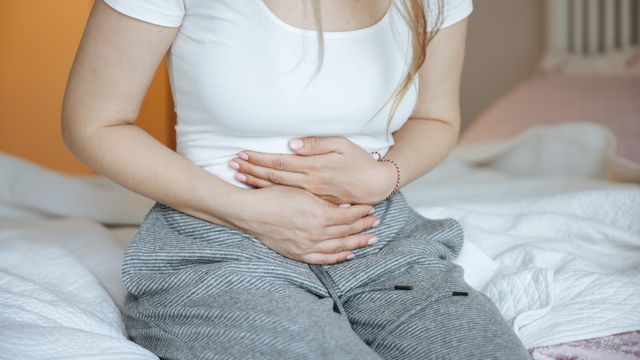 A woman suffering from stomach pain sits on a bed holding her stomach. 