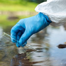 Gloved hand collecting a water sample in a test tube from a river. 