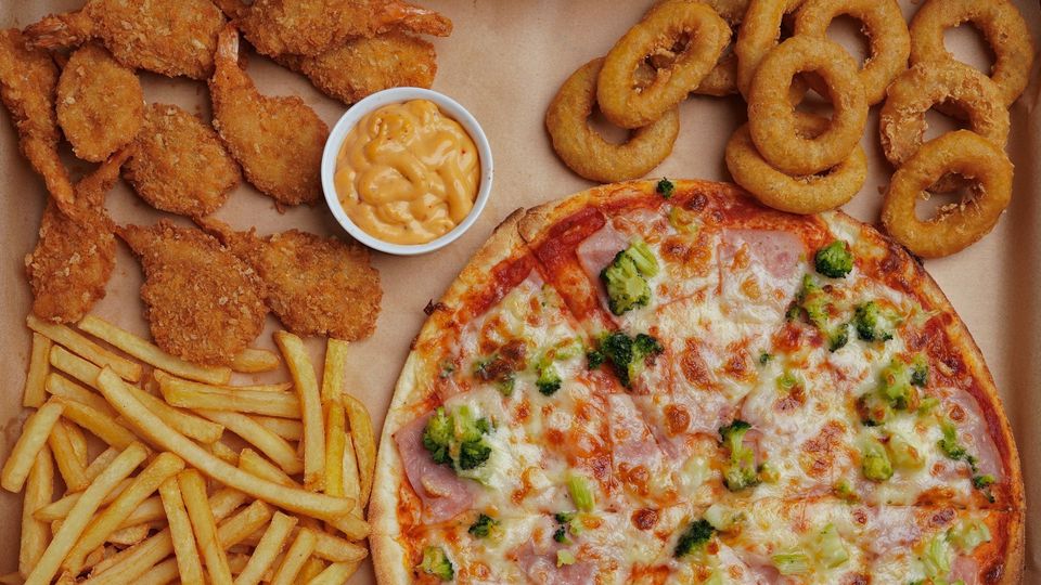 Flat-lay photograph of fatty foods including pizza, fries, onion rings, and battered prawns.