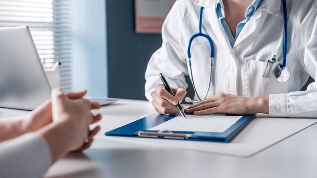 A doctor writing notes on paper while sitting opposite their patient in a clinical setting. 
