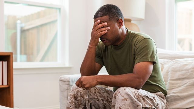 Person in military-style camouflage trousers seated on a sofa with hand covering face, representing PTSD. 