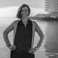 Woman researcher standing confidently by a waterfront with modern buildings in the background.
