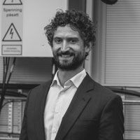 A black and white image of Marius, smiling at the camera. He has curly hair and a beard, and is wearing a dark-colored suit.